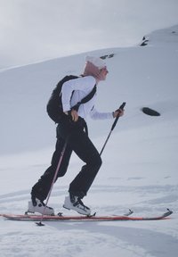Skieur portant des bottes blanches et un pantalon noir qui remonte une pente enneigée, tenant deux bâtons de ski, avec un sac à dos noir et un tour de cou rose.