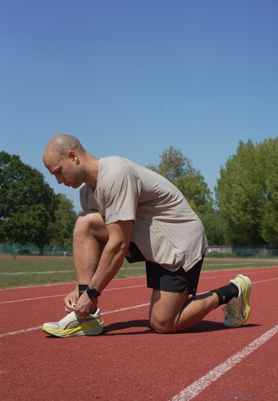 Hombre arrodillado en una pista de atletismo roja al aire libre, atándose las zapatillas blancas para correr, vistiendo pantalones cortos negros y una camisa de color claro bajo un cielo azul despejado.