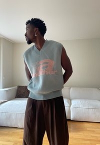 Mann mit Ohrringen, der eine ärmellose Strickweste und weite braune Hosen trägt, steht in einem Wohnzimmer mit einem weißen Sofa.