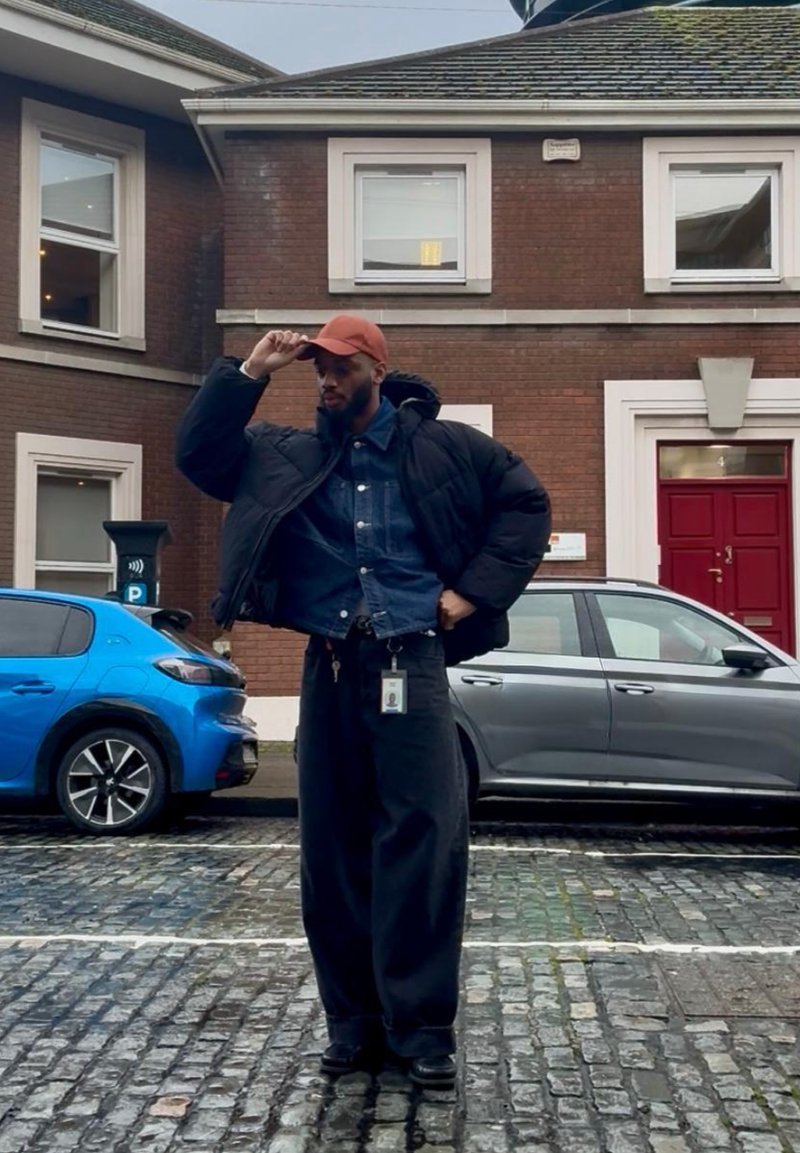 Homme portant une veste matelassée noire, une chemise en denim et une casquette orange, debout sur une rue pavée à côté de voitures bleues et grises devant un bâtiment en briques.