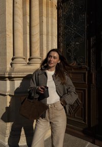 Hnedé kárované sako cez svetlý sveter, béžové vysoký pás nohavice a hnedá semišová taška. Drží čierny pohár na kávu, stojí pri zdobných dverách.