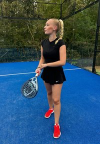 Černý sportovní top a sukně, červené sportovní boty, držící pádlo s otvory. Modrý povrch kurtu a zelené listoví v pozadí.