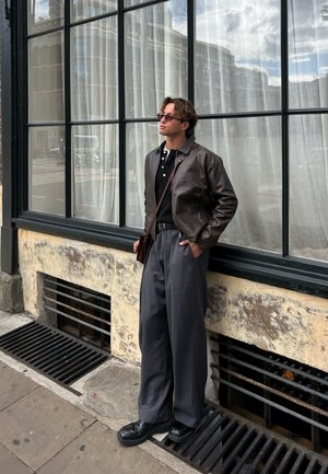 Chaqueta de cuero marrón, camisa polo negra, pantalones de pierna ancha grises, zapatos negros, de pie junto a una ventana con cortinas translúcidas. Entorno urbano al aire libre.