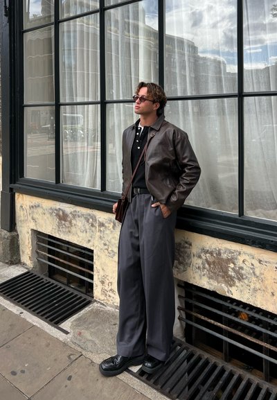 Chaqueta de cuero marrón, camisa polo negra, pantalones de pierna ancha grises, zapatos negros, de pie junto a una ventana con cortinas translúcidas. Entorno urbano al aire libre.