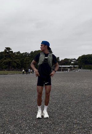 Hombre atlético con un chaleco verde claro sobre una camiseta negra, pantalones cortos negros con un logo blanco de Nike y zapatillas blancas, de pie al aire libre.