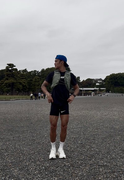 Hombre atlético con un chaleco verde claro sobre una camiseta negra, pantalones cortos negros con un logo blanco de Nike y zapatillas blancas, de pie al aire libre.