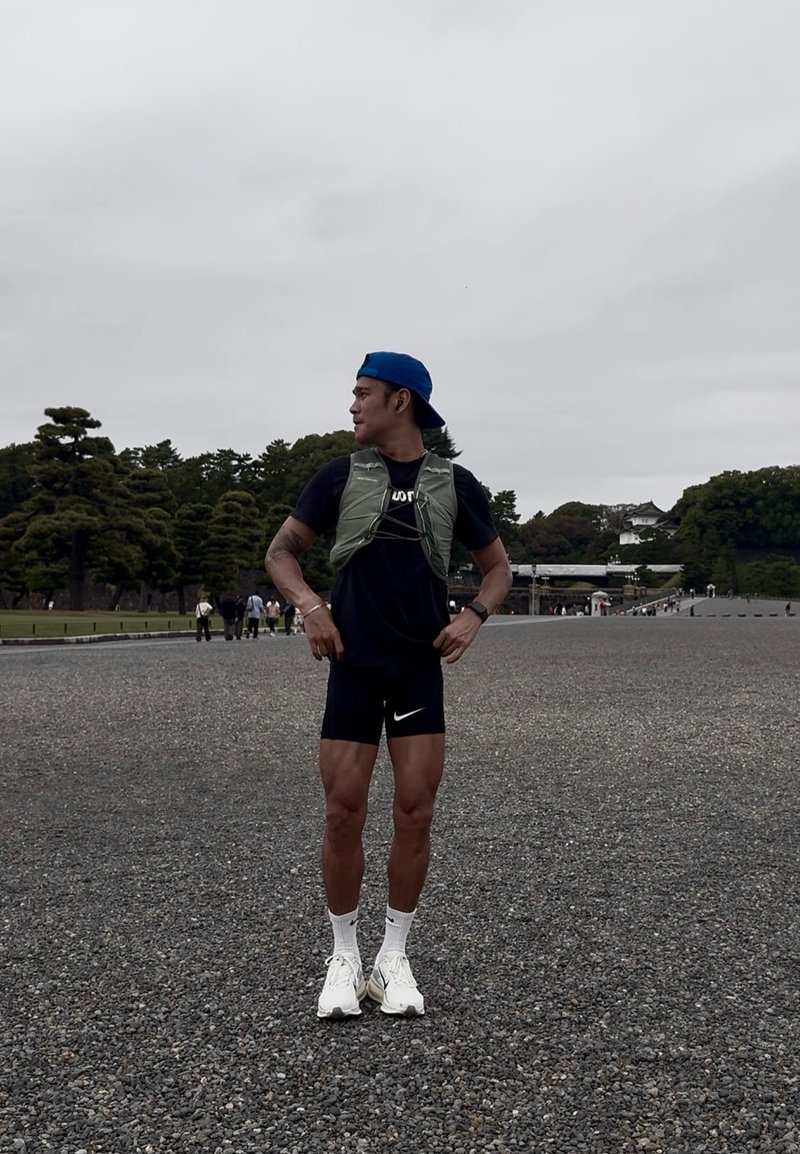 Uomo atletico con canotta verde chiaro sopra una maglia nera, pantaloncini neri con logo Nike bianco e scarpe da ginnastica bianche, in piedi all'aperto.