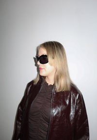 Femme aux cheveux blonds portant de grandes lunettes de soleil noires, un haut foncé froncé et une veste en cuir rouge foncé brillante sur un fond blanc uni.