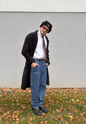 Abrigo largo negro, camisa blanca, corbata marrón, vaqueros holgados azules, zapatos negros y gafas de sol redondas naranjas, llevando un gorro negro.