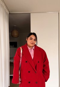 Donna con capelli scuri che indossa un cappotto rosso e una maglietta rosa si trova all'interno vicino a una porta scorrevole con una borsa a tracolla sulla spalla sinistra.