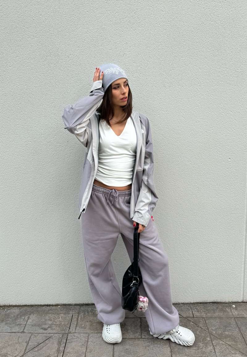 Jeune femme avec bonnet gris, veste légère, haut blanc, pantalon de jogging gris et baskets blanches, debout contre un mur uni tenant un petit sac noir.