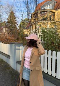 Manteau long brun clair, cardigan côtelé rose pastel, jupe en denim, et casquette rose. Entourée d'arbres et d'une clôture en bois blanc.