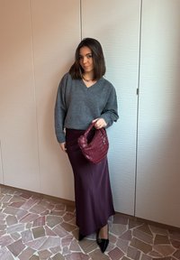 Μια γυναίκα στέκεται μέσα σε εσωτερικό χώρο φορώντας γκρι πουλόβερ και μακριά μωβ φούστα, κρατώντας μια πλεκτή μπορντό τσάντα, με μαύρα μυτερά παπούτσια πάνω σε ψηφιδωτό πάτωμα.