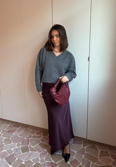 Mujer en interiores con suéter gris y falda larga morada, sosteniendo un bolso tejido color burdeos, con zapatos negros puntiagudos sobre suelo de mosaico.