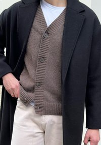 Person in einem schwarzen Mantel über einem braunen, geknöpften Cardigan und einem weißen Hemd, mit hellfarbener Hose und einer Hand in der Tasche.