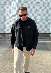 Melna pogājama krekls ar divām krūšu kabatām, kopā ar melnu T-kreklu un krēmkrāsas biksēm. Vīrietis nēsā saulesbrilles un staigā pa betonu.