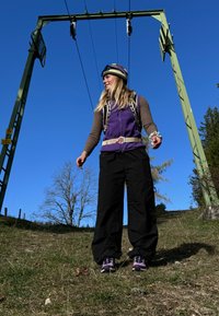 Lila polár mellény barna hosszú ujjú ing felett, fekete bő nadrág és lila sportcipő. Hord magán egy hevedert és pántokat, kék ég alatt.