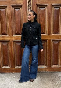 Svart tettsittende jakke med sølvknapper og fire lommer, kombinert med vide blå denimjeans og brune hæler, foran en trebakgrunn.