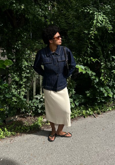 Chaqueta de mezclilla oscura con dos bolsillos en el pecho, combinada con una falda midi crema y chanclas negras. La vegetación circundante ofrece un fondo natural.