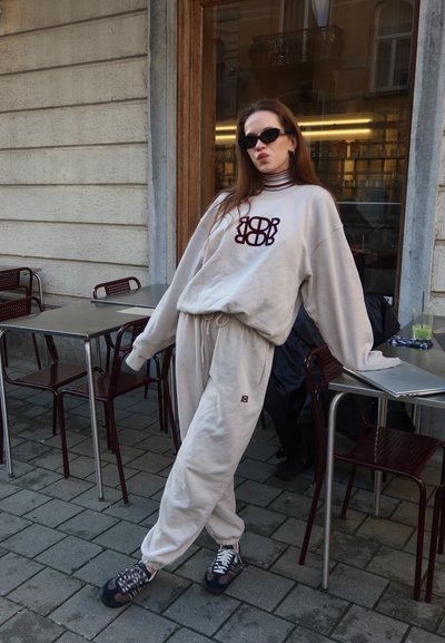 Sudadera y pantalones jogger de color beige claro con un logo impreso. Tejido texturizado, corte holgado, combinados con zapatillas negras y gafas de sol. Entorno de café al aire libre.