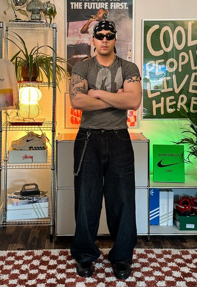 Pantalones de mezclilla oscuros de pierna ancha con un detalle de cadena, camiseta gráfica gris, gafas de sol negras y zapatillas negras. El fondo presenta plantas y carteles.