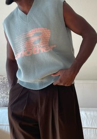 Mann trägt ärmellose hellblaue Strickweste mit rosa Grafiktext und dunkelbraune plissierte Hose, Hand ruht auf der Hüfte drinnen.