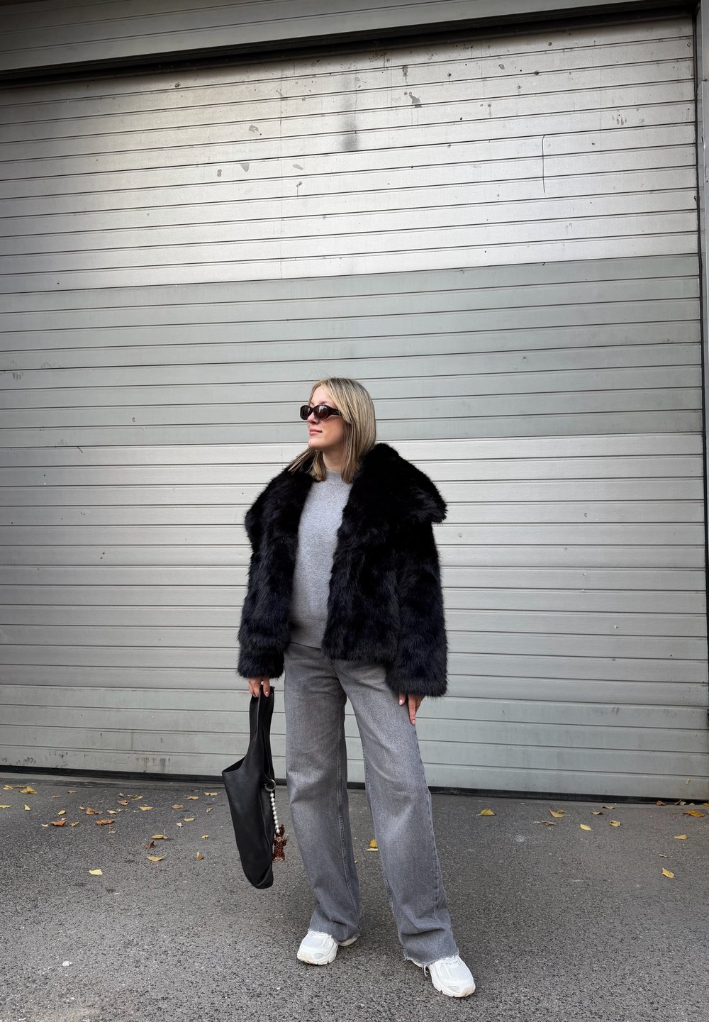 Casaco de pelo sintético preto com colarinho largo, suéter cinza por baixo, calças largas cinzas e mala tote preta. Ténis complementam o look.