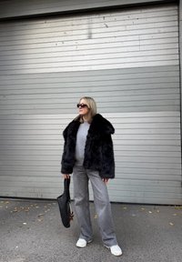 Casaco de pelo sintético preto com colarinho largo, suéter cinza por baixo, calças largas cinzas e mala tote preta. Ténis complementam o look.