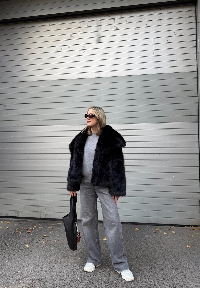 Chaqueta de piel sintética negra con cuello amplio, suéter gris debajo, pantalones grises de pierna ancha y bolso tote negro. Zapatillas completan el conjunto.