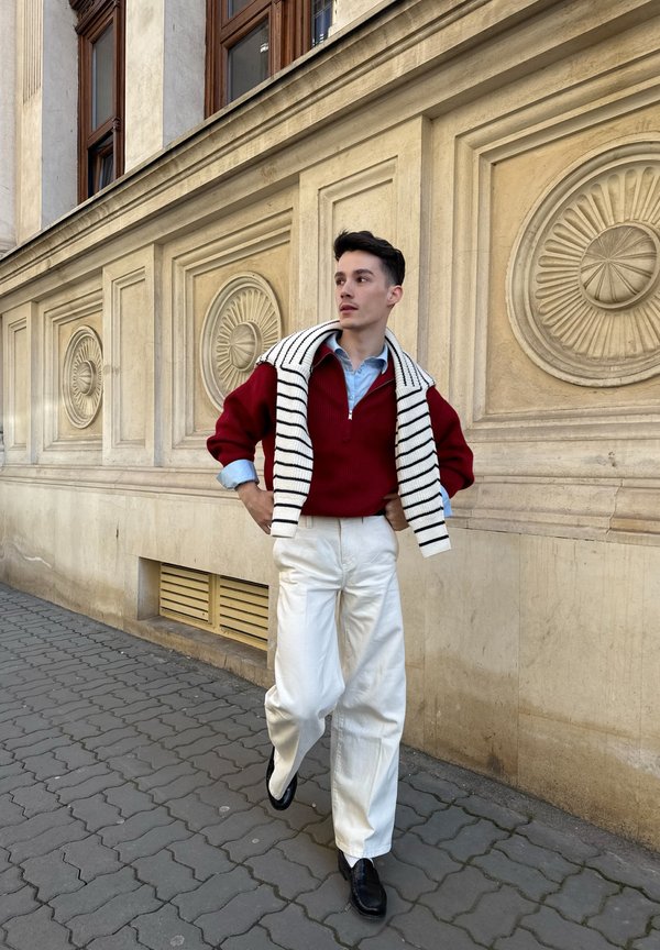 Jonge man wandelt op de stoep in de stad, draagt witte broek, rode trui, lichtblauw hemd en gestreepte sjaal over de schouders.
