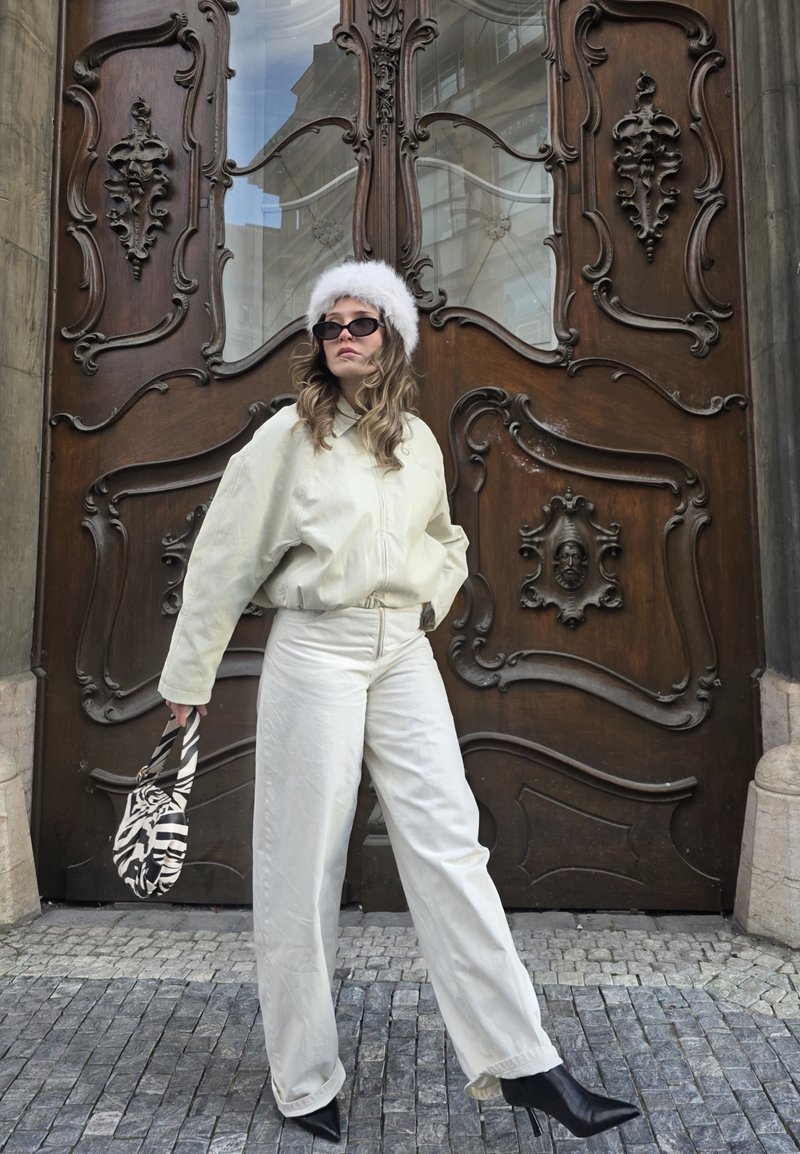 Giacca e pantaloni oversize color crema, abbinati a un cappello di pelliccia bianca e stivali neri con tacchi alti. Tiene in mano una borsa con motivo a zebra.