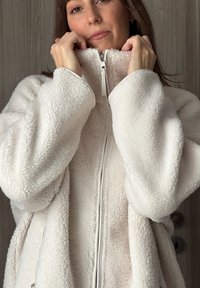 Γυναίκα με ανοιχτόχρωμο δέρμα και καστανά μαλλιά κλειδώνει το φερμουάρ ενός λευκού, υφασμάτινου fleece, στέκεται μπροστά σε ένα ξύλινο φόντο πόρτας.