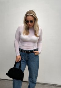 Femme blonde portant des lunettes de soleil, un haut transparent blanc à manches longues, un jean bleu avec une ceinture noire, tenant un petit sac à main noir, debout contre un mur gris.