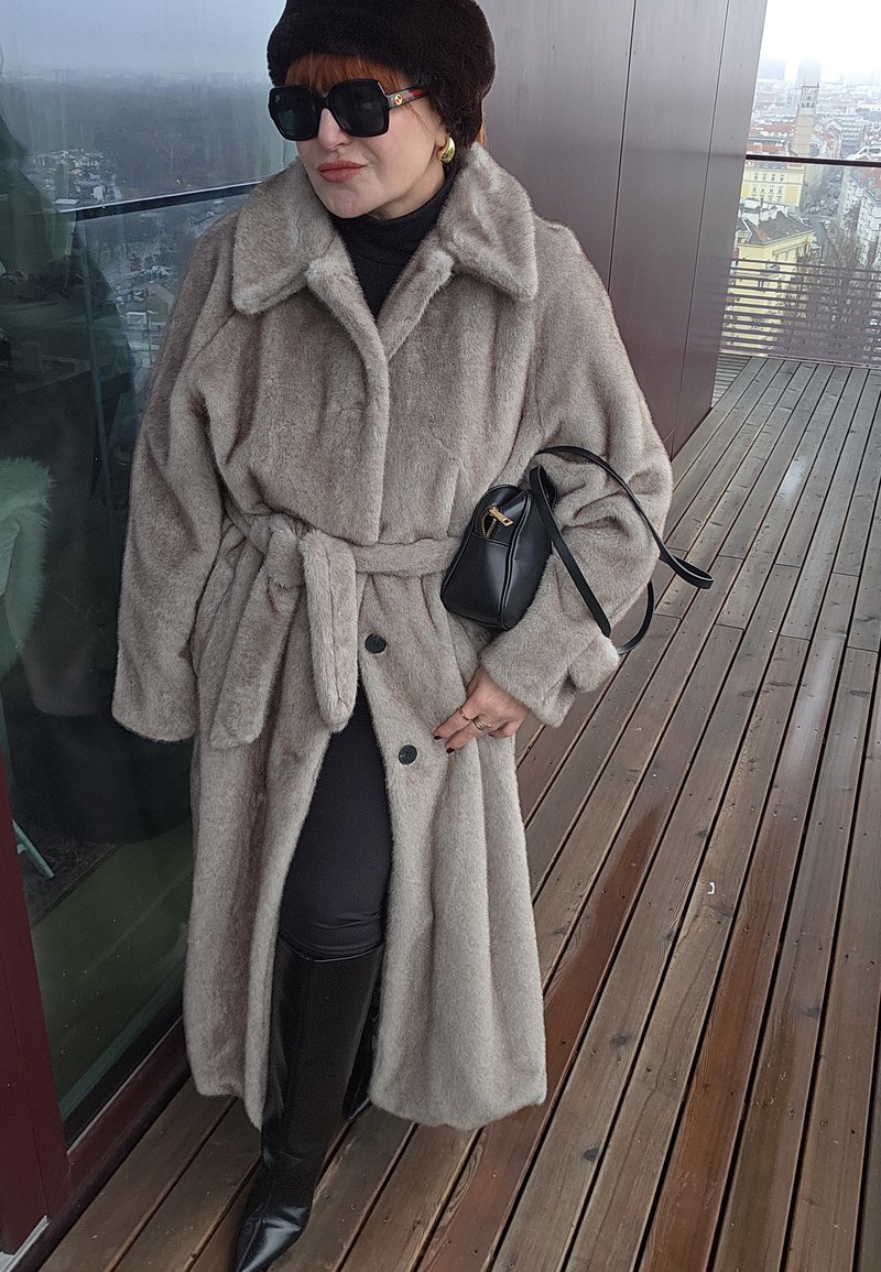 Manteau en fausse fourrure beige clair avec un large col, une ceinture nouée à la taille et de gros boutons. Porté avec des leggings noirs et des bottes hautes jusqu'aux genoux.