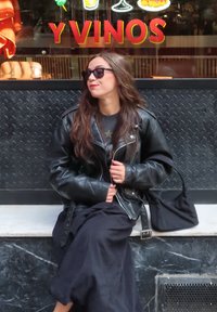 Svart skinnjakke med sølvfargede detaljer, lagd over en mørkegrå skjorte. Lang svart skjørt og en liten svart veske. Mørke briller, uformell pose.