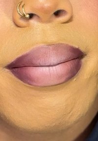 Ingrandimento delle labbra con un rossetto matte rosa e un sottile effetto ombre. La pelle ha un colore abbronzato con una texture visibile. Anello da naso a cerchio dorato.