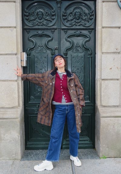 Abrigo marrón de cuadros de gran tamaño sobre una camisa de botones burdeos y una camiseta a rayas, combinado con jeans de mezclilla y zapatillas blancas, frente a una puerta verde detallada.