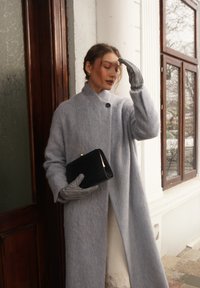 Γυναίκα με μακρύ γκρι παλτό και γάντια που ταιριάζουν κρατά ένα μαύρο πορτοφόλι, στέκεται κοντά σε μια ξύλινη πόρτα και παράθυρο με θέα στο χειμωνιάτικο εξωτερικό τοπίο.