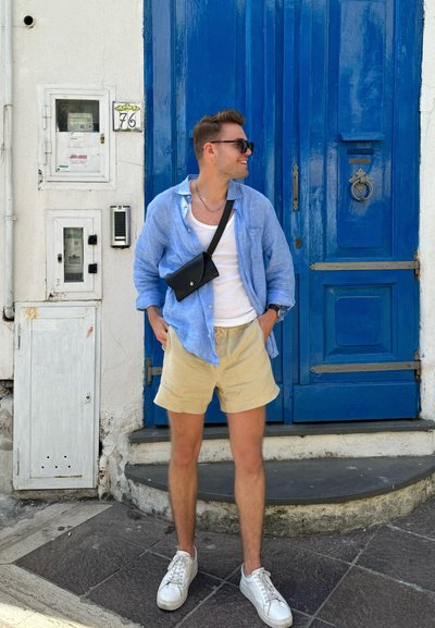 Camisa de lino azul claro, camiseta sin mangas blanca, shorts beige, bolso cruzado negro y zapatillas blancas, con una puerta azul brillante de fondo.