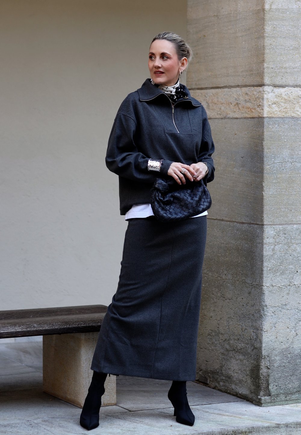 Femme portant une longue jupe grise foncée, un pull assorti, des bottes noires, une écharpe à pois, des bijoux argentés, tenant une pochette noire tissée.