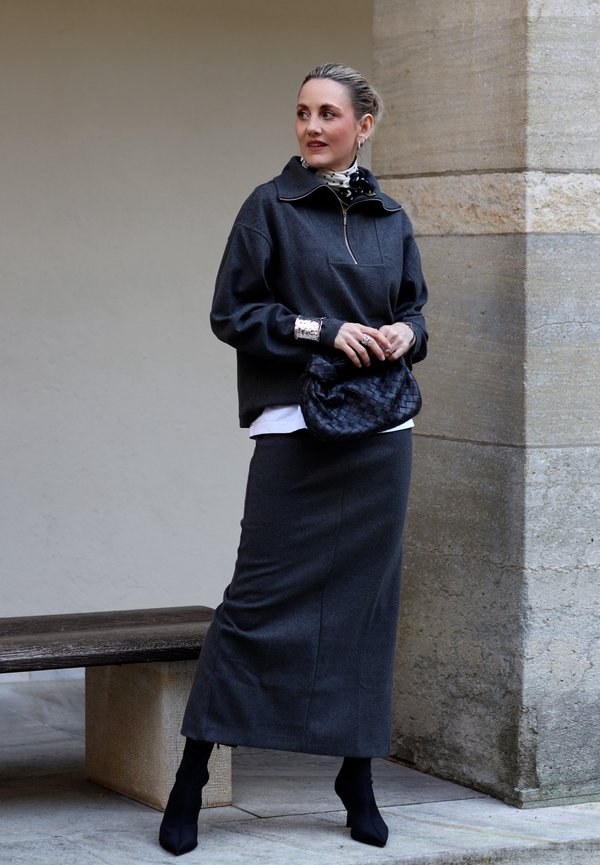 Frau trägt einen dunkelgrauen langen Rock, passenden Pullover, schwarze Stiefel, gepunkteten Schal, silbernen Schmuck und hält eine schwarze gewebte Clutch-Tasche.