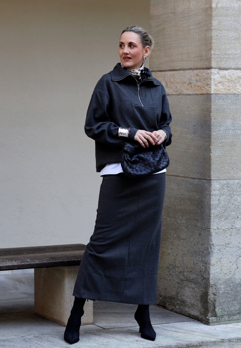 Donna che indossa una gonna lunga grigio scuro, pullover abbinato, stivali neri, sciarpa a pois, gioielli d'argento e tiene in mano una pochette nera intrecciata.