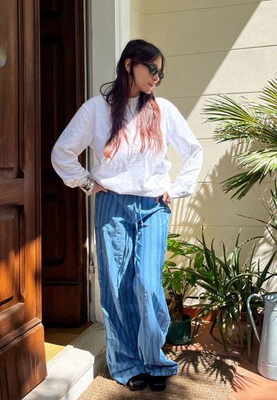 Camisa blanca de manga larga con un corte relajado, pantalones holgados azules con rayas verticales y zapatos negros. Plantas verdes al fondo y una puerta de madera.