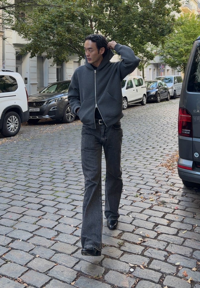 Homme en sweat à capuche sombre et jean marchant sur une rue pavée bordée de voitures garées et d’arbres dans un quartier résidentiel urbain.