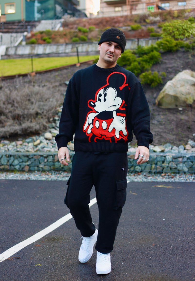Férfi sétál kint, fekete sapkát, fekete Mickey egér mintás pulóvert, fekete cargo nadrágot és fehér tornacipőt visel egy burkolt úton.