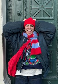 Jeune personne portant un bonnet rouge, une écharpe rouge et bleue, une veste noire et un sac rouge, riant les mains sur la tête devant une porte ornée vert foncé.