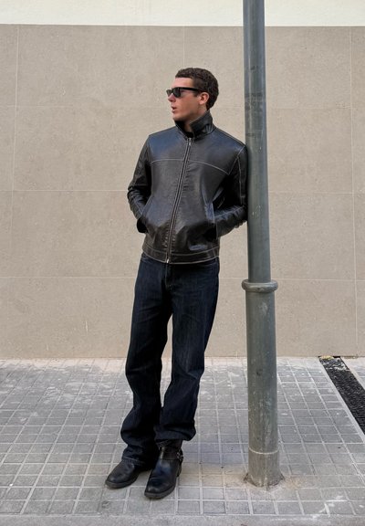 Hombre con chaqueta de cuero negra, vaqueros oscuros y botas apoyado contra un poste de metal en una acera de baldosas grises con fondo de pared beige.