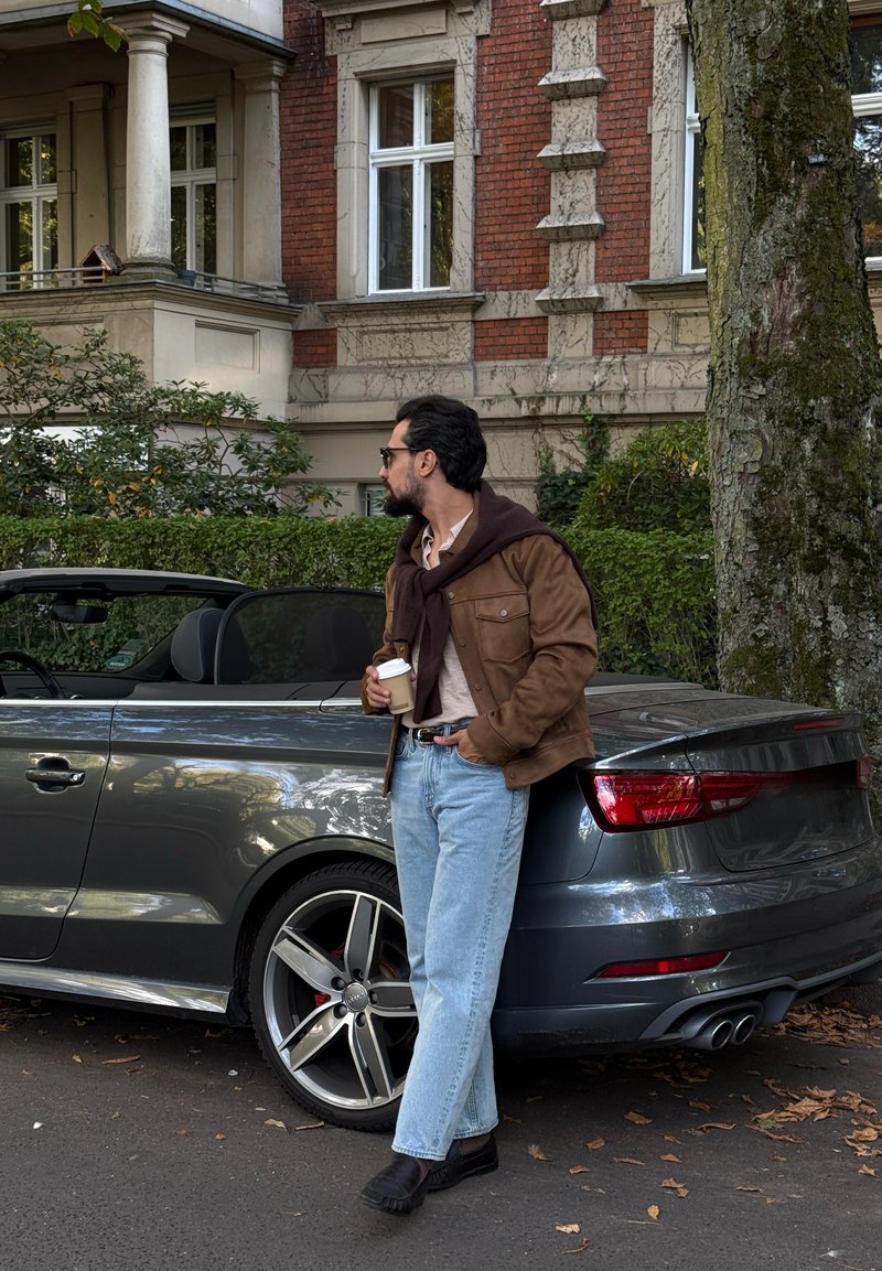 Homme portant des lunettes de soleil, une veste marron et un jean bleu s'appuie sur une voiture décapotable grise en tenant une tasse de café dans une rue avec des arbres et des bâtiments en brique.