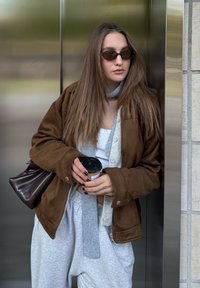 Bruine suède jas over een witte crop top, grijze sweatpants, grijze sjaal, zwarte zonnebril, houdt een koffiekop vast, donkere tas. Metalen achtergrond.