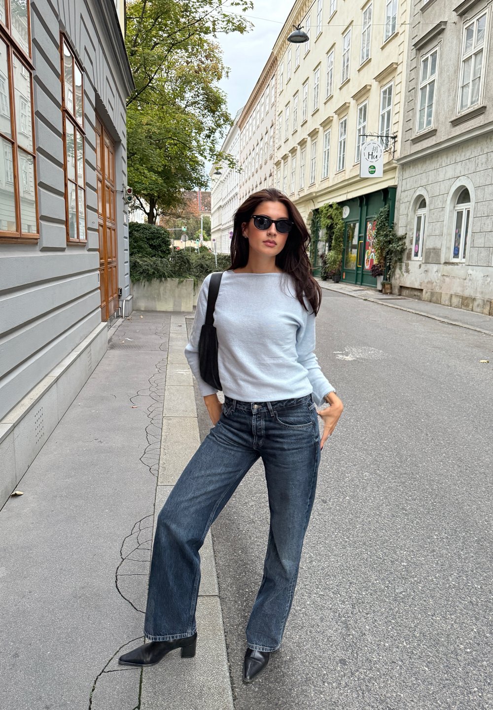 Maglione a maniche lunghe di un blu chiaro, jeans a gamba larga scuri, stivali neri a punta e occhiali da sole neri, in piedi su una strada cittadina.
